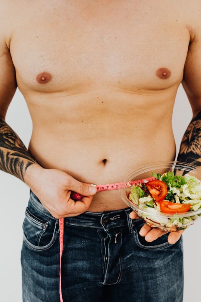 Tattooed man measuring waist while holding a salad bowl for healthy lifestyle and weight loss concept.